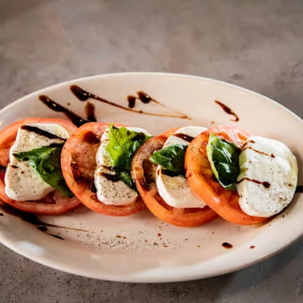 A Caprese salad with sliced tomatoes, mozzarella, basil, and balsamic glaze on a white plate.