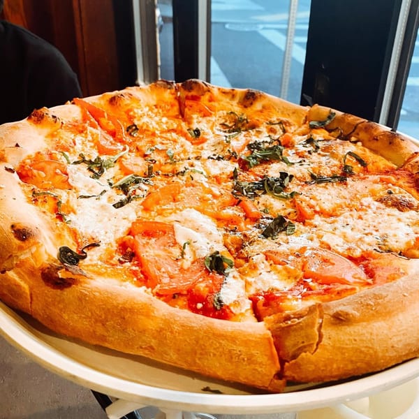 A large, freshly baked Rocco's pizza with tomatoes, cheese, and basil on a white stand.