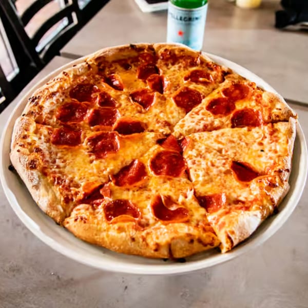 A whole pepperoni pizza with several slices cut, on a white plate, with a bottle in the background.