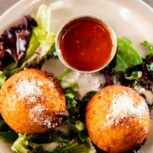 Two golden-brown arancini balls dusted with parmesan cheese, served with marinara sauce and greens.